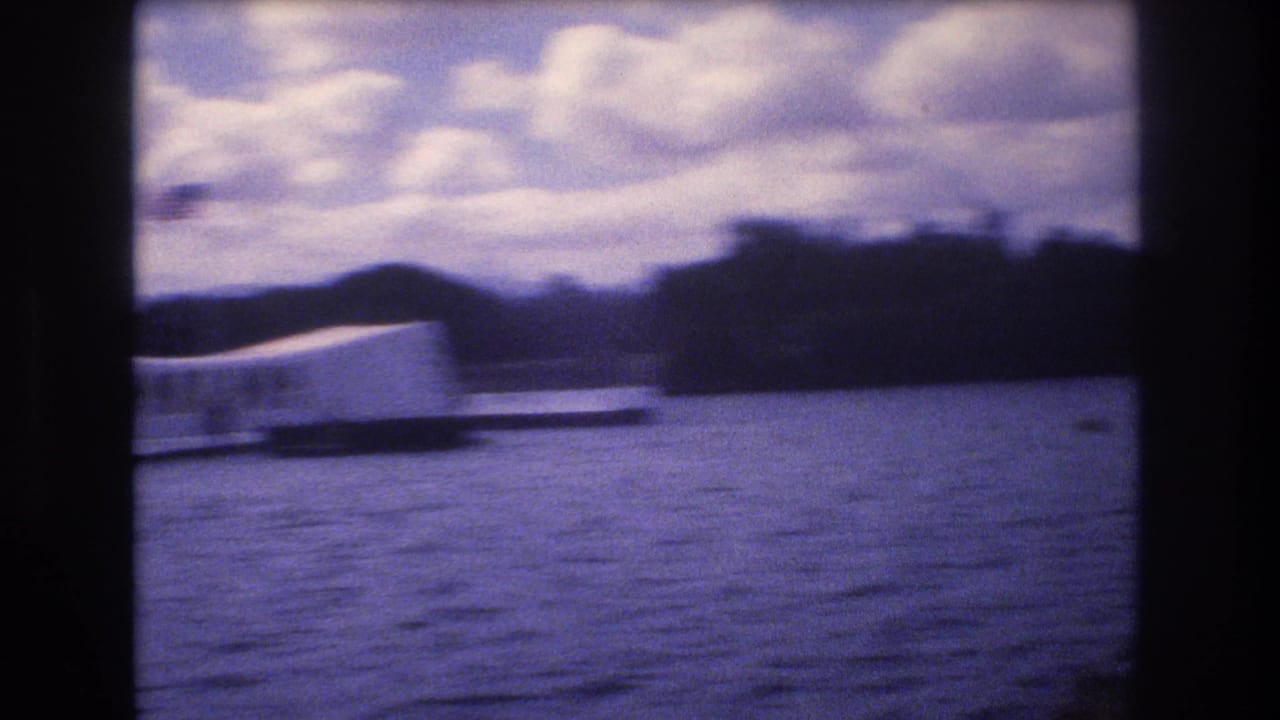 1977 Honolulu home-movie frame showing the USS Arizona Memorial across Pearl Harbor water, partly obscured by dark boat supports and a purple 8mm color cast.