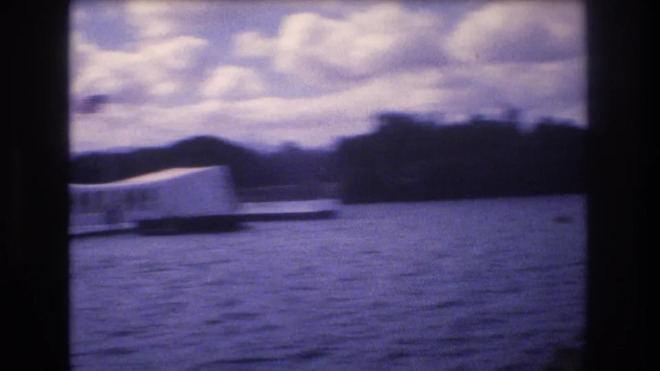 1977 Honolulu home-movie frame showing the USS Arizona Memorial across Pearl Harbor water, partly obscured by dark boat supports and a purple 8mm color cast.