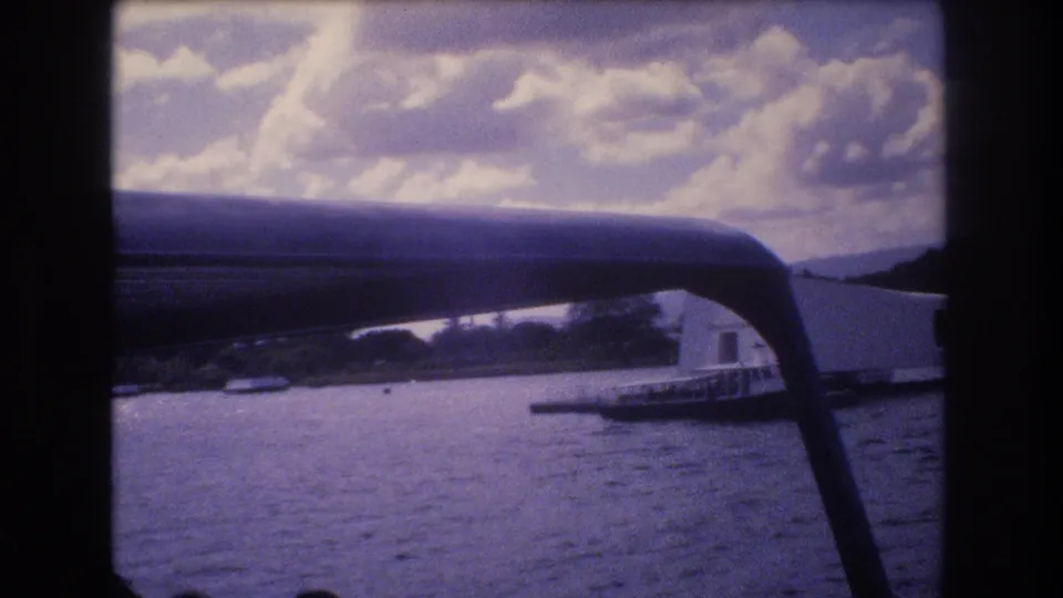 Home-movie frame from Pearl Harbor in 1977 showing the white USS Arizona Memorial beyond harbor water, framed by a curved boat support and faint visitor silhouettes.