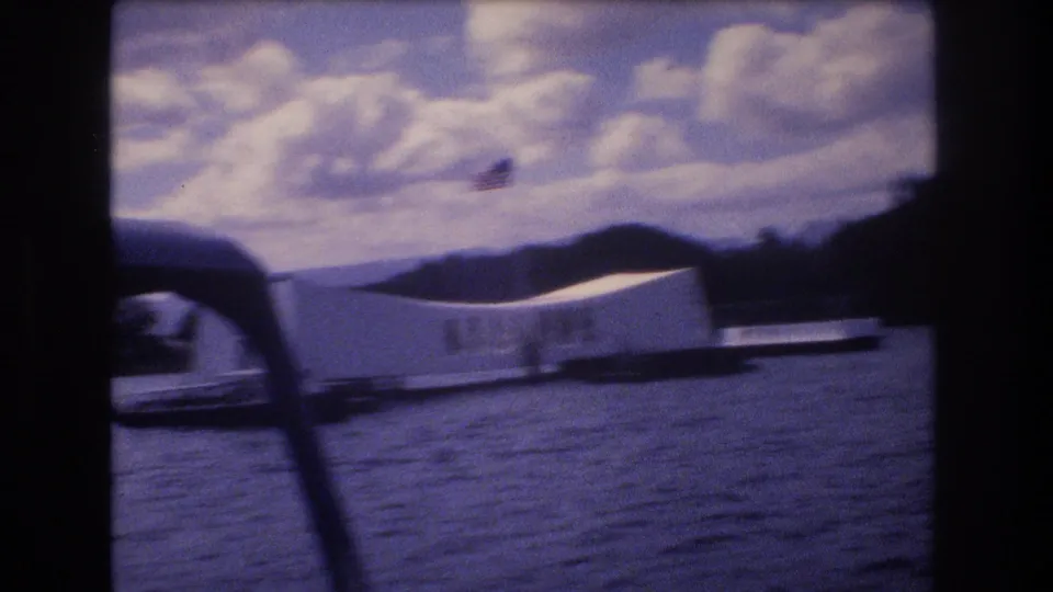1977 8mm frame of the USS Arizona Memorial at Pearl Harbor, viewed from a moving boat with dark foreground supports, rippled water, and cloudy sky.
