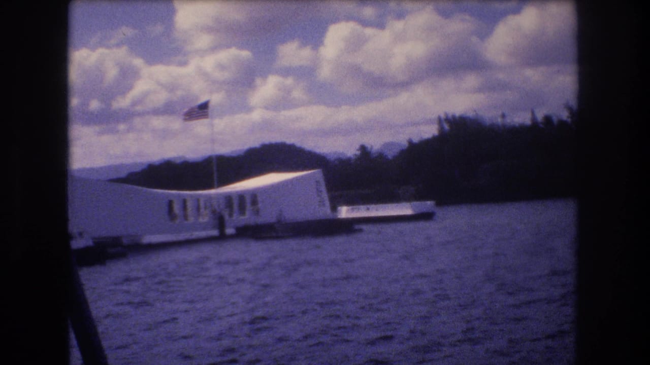 1977 8mm home-movie frame from Honolulu showing the USS Arizona Memorial viewed from a boat, with harbor water, an American flag, distant hills, and dark film borders.
