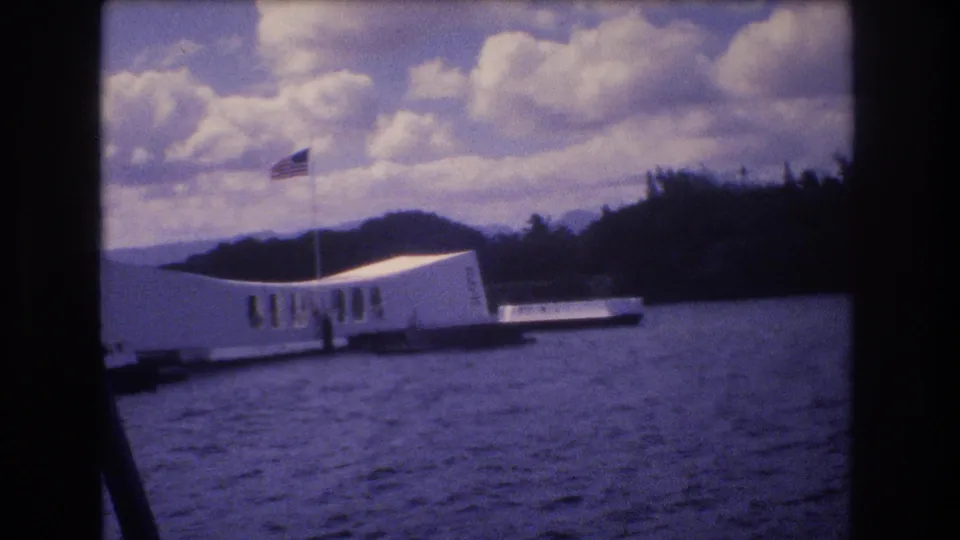 1977 8mm home-movie frame from Honolulu showing the USS Arizona Memorial viewed from a boat, with harbor water, an American flag, distant hills, and dark film borders.