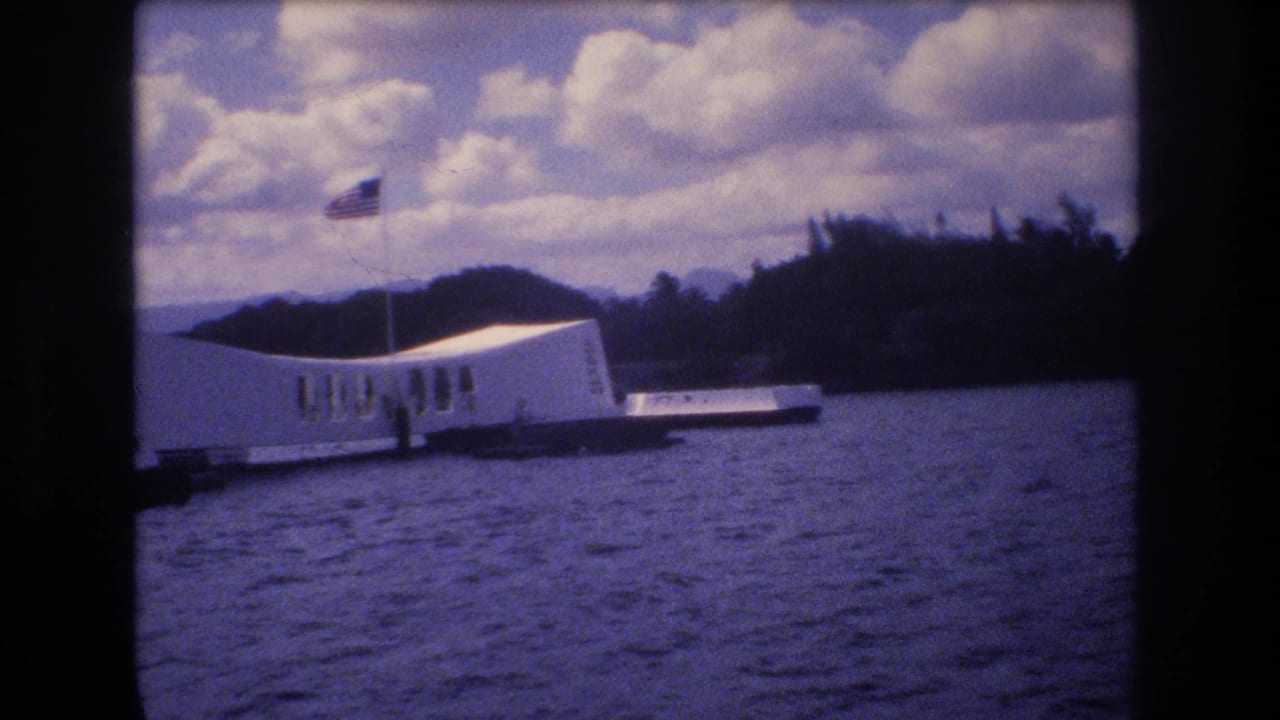 White memorial structure on the water with a flagpole, clouds, and hills behind it.
