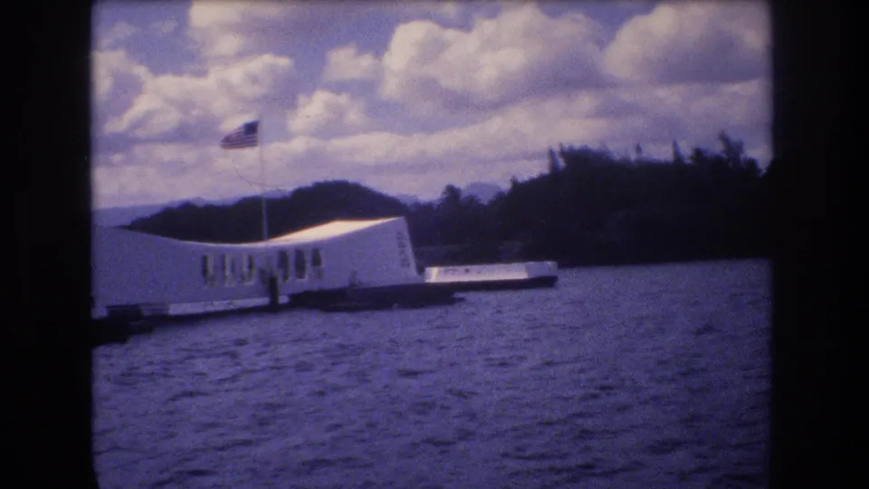 The USS Arizona Memorial appears from a boat-level viewpoint in Honolulu, with its white structure crossing the harbor view beneath a cloudy sky and American flag.