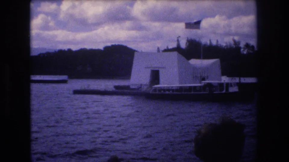 1977 Honolulu home-movie frame showing a tour boat moving in front of the USS Arizona Memorial, with harbor water, distant hills, cloudy sky, and a dark foreground edge.