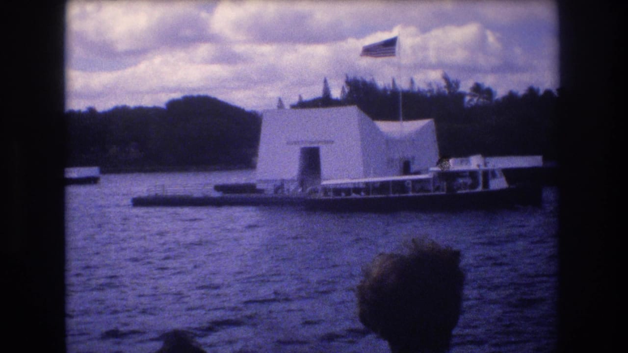 1977 8mm home-movie frame of the USS Arizona Memorial in Honolulu viewed from the water, with a tour boat nearby, an American flag above the structure, and a foreground passenger silhouette.