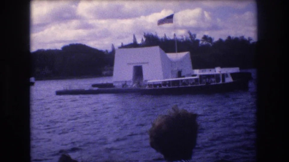 1977 Honolulu harbor view of the USS Arizona Memorial from a moving vessel, showing the white structure, flag, nearby tour boat, rippled water, and passenger silhouette at the bottom edge.