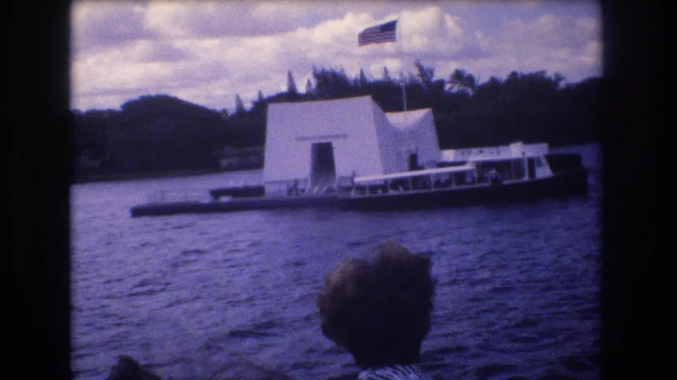 1977 8mm frame showing the USS Arizona Memorial and a low tour boat on Honolulu harbor, with cloudy sky, shoreline trees, and a foreground visitor silhouette from the filming boat.