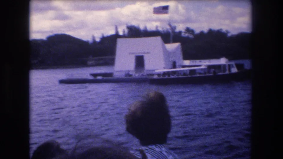 Blue-purple 8mm home-movie frame from 1977 showing the USS Arizona Memorial in Honolulu from the water, with the American flag visible above and a dark visitor silhouette in the foreground.