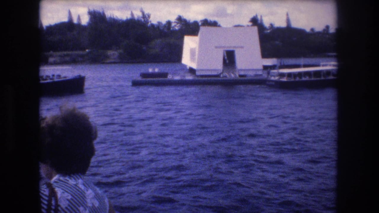 1977 Honolulu home-movie frame showing the USS Arizona Memorial across the water, with tour boats nearby and a visitor seen from behind in the foreground.