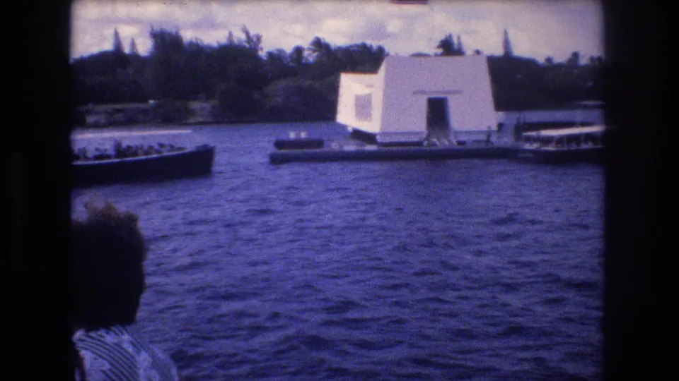 The USS Arizona Memorial appears across the harbor in Honolulu, with small tour boats nearby and a visitor partially visible along the left edge.