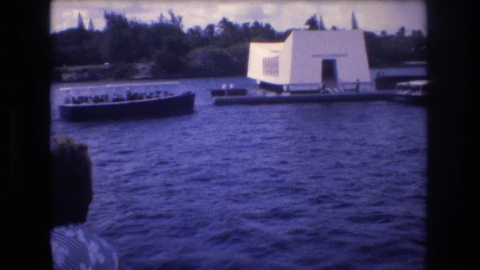 A 1977 Honolulu frame shows a passenger-filled tour boat near the USS Arizona Memorial, with another visitor visible from behind in the lower left.