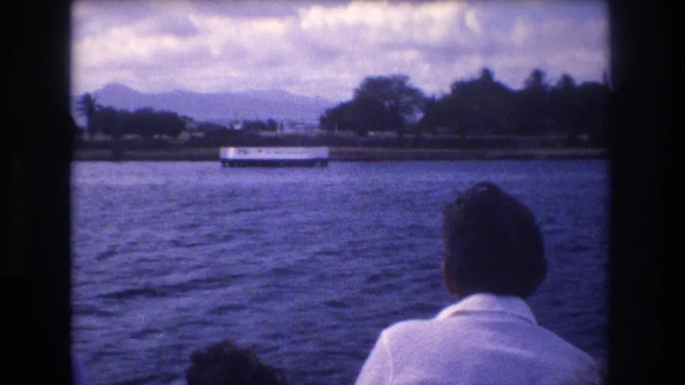 The camera turns across Honolulu harbor toward a distant boat, shoreline trees, cloudy sky, and mountains, with a visitor seen from behind in the foreground.
