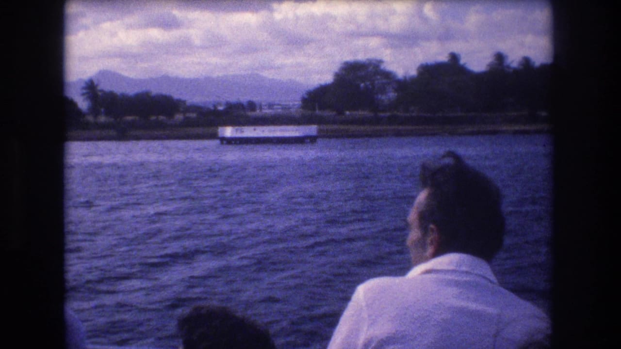 1977 Honolulu home-movie frame showing a man in a white shirt seated on a moving boat, looking over rippling water toward shoreline trees, a white structure, and distant mountains.