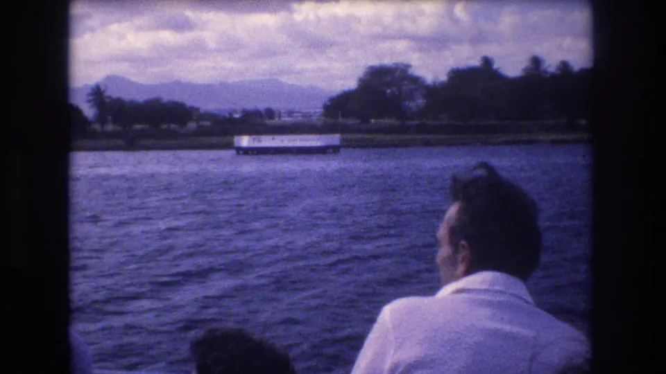 1977 Honolulu home-movie frame showing a man in a white shirt seated on a moving boat, looking over rippling water toward shoreline trees, a white structure, and distant mountains.