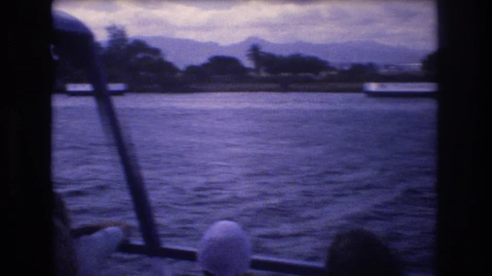 1977 Honolulu boat-view frame with dark window supports in the foreground, rippling water across the middle ground, and a low shoreline with trees and mountains under cloudy sky.