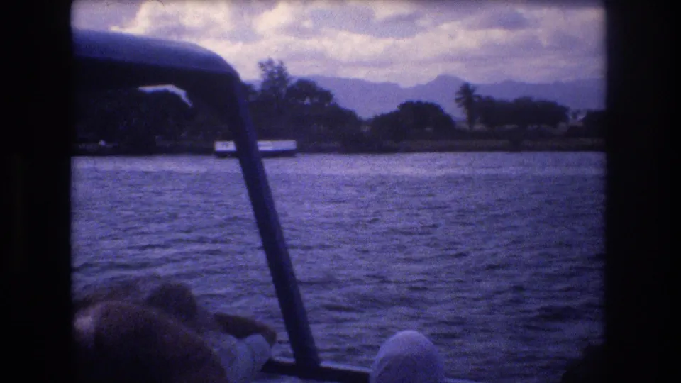 1977 Honolulu home-movie frame looking from a moving boat across harbor water, with a partial passenger at the lower edge, a dark boat support, shoreline vegetation, and hazy mountains beyond.