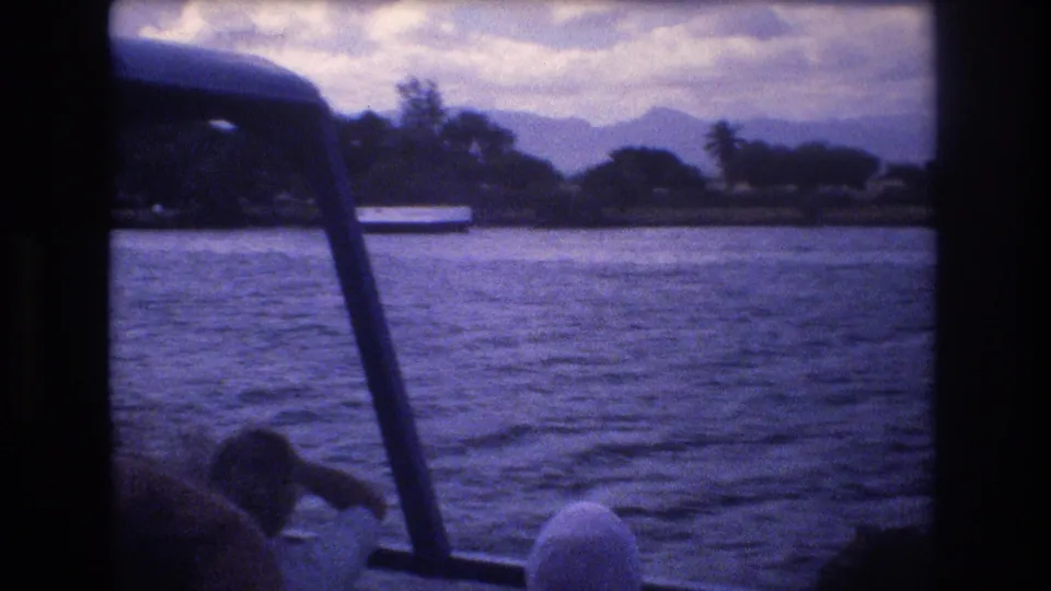 1977 Honolulu home-movie frame showing a partial passenger near the lower edge of the boat view, rippling harbor water, a white shoreline structure, trees, and mountains under a cloudy sky.