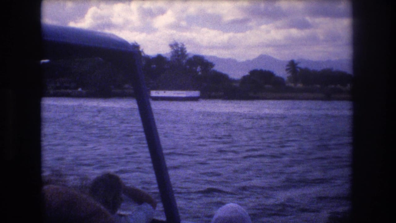1977 Honolulu 8mm boat-view frame showing rippling water, a tropical shoreline with palm trees, distant mountains, and passengers partially visible in the foreground.