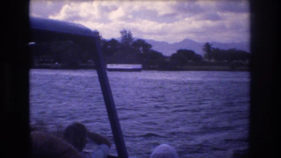 1977 Honolulu 8mm boat-view frame showing rippling water, a tropical shoreline with palm trees, distant mountains, and passengers partially visible in the foreground.