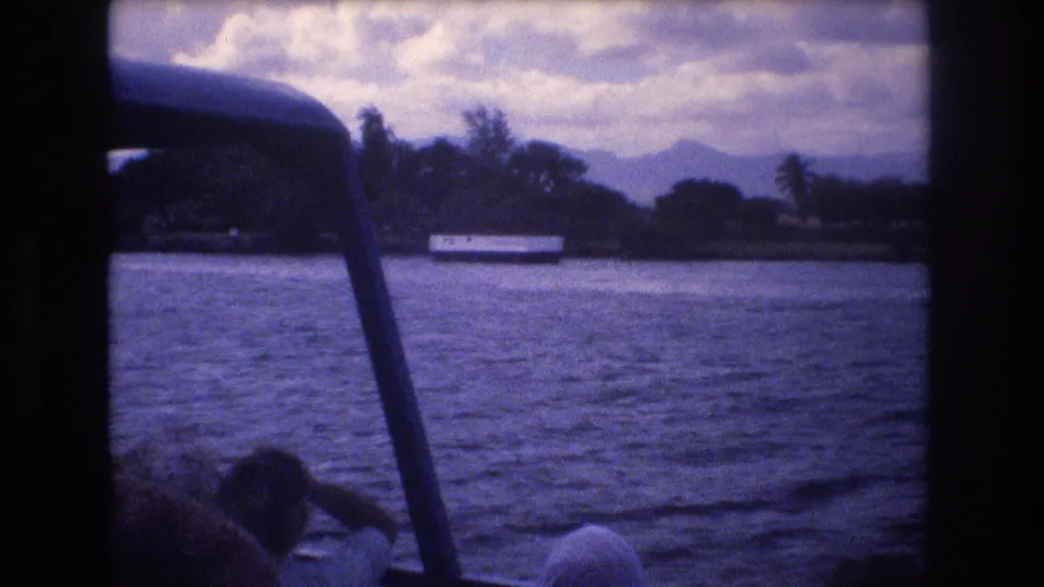 1977 Honolulu home-movie frame from a moving boat, with passengers at the bottom edge, open water, shoreline trees, and mountains under a cloudy sky.