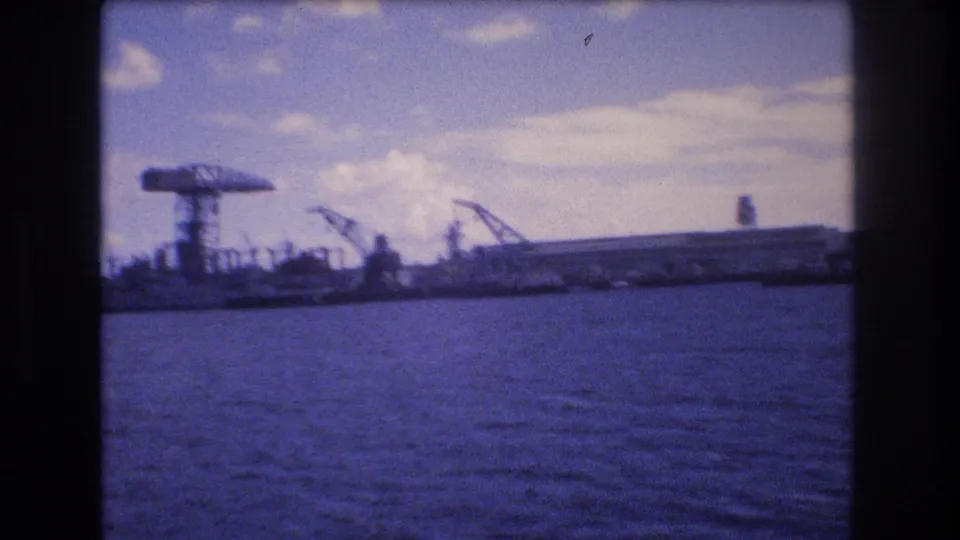 1977 Honolulu 8mm frame showing an industrial waterfront from the water, with large cranes, low port structures, harbor surface, and partly cloudy sky.
