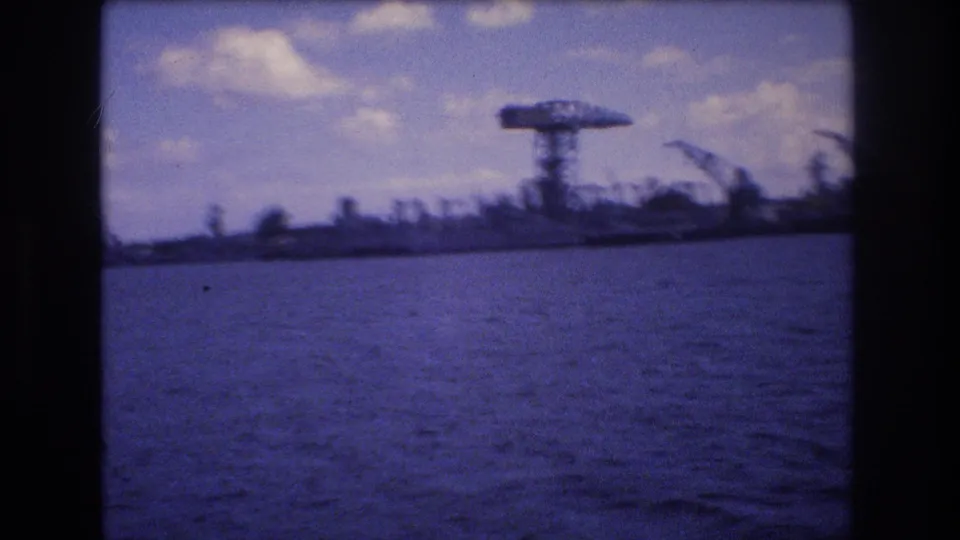 1977 Honolulu home-movie frame looking across harbor water toward cranes and port structures, with a bright partly cloudy sky and soft 8mm color cast.