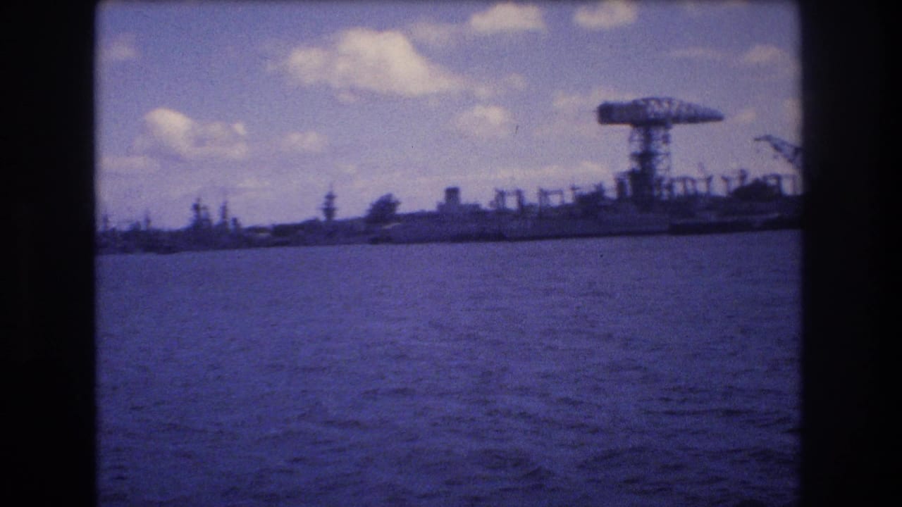 1977 Honolulu harbor view from the water, showing rippling foreground water, distant industrial cranes, and large docked vessels under scattered clouds.