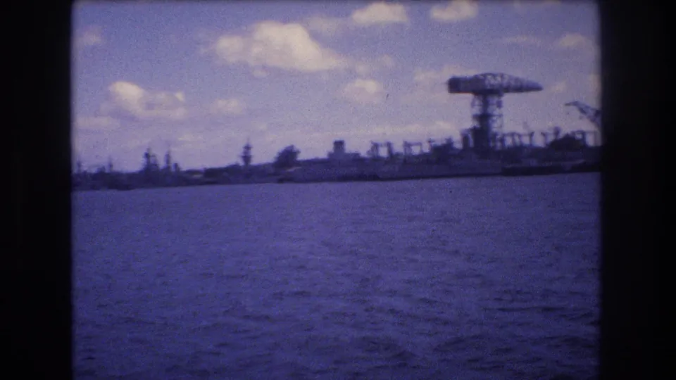 1977 Honolulu harbor view from the water, showing rippling foreground water, distant industrial cranes, and large docked vessels under scattered clouds.
