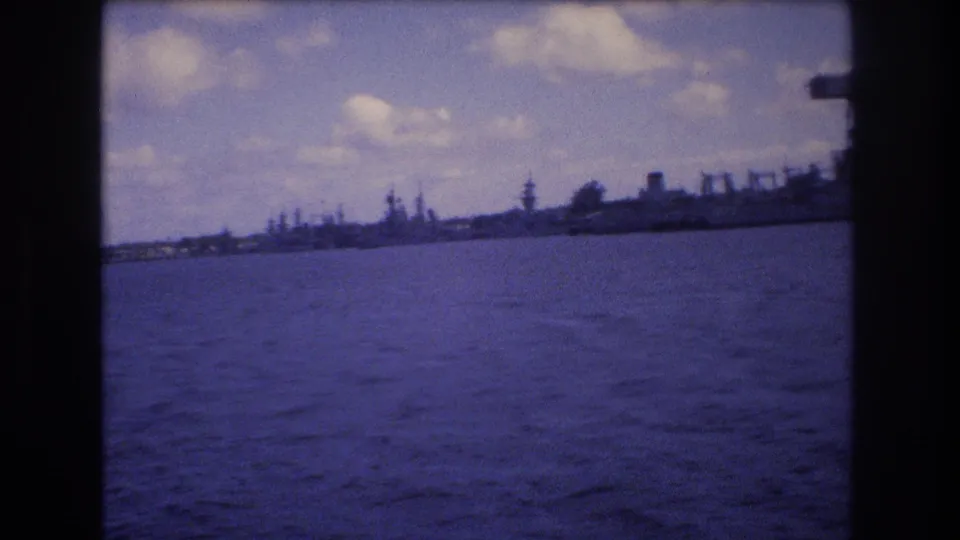 1977 Honolulu water-level view toward a distant harbor shoreline with cranes, low dock structures, and large vessels softened by 8mm film grain.