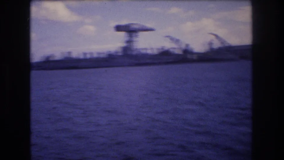 Motion-blurred 1977 Honolulu harbor frame from a moving boat, with blue water, a prominent dockside crane, low industrial structures, and clouds.