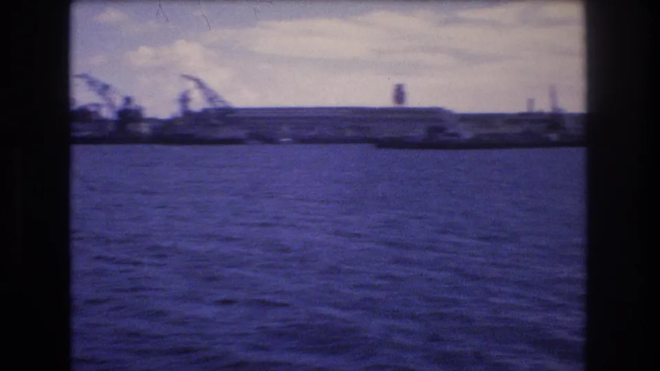 1977 Honolulu harbor view across open water toward a low industrial waterfront with cranes and distant vertical structures under a bright cloudy sky.