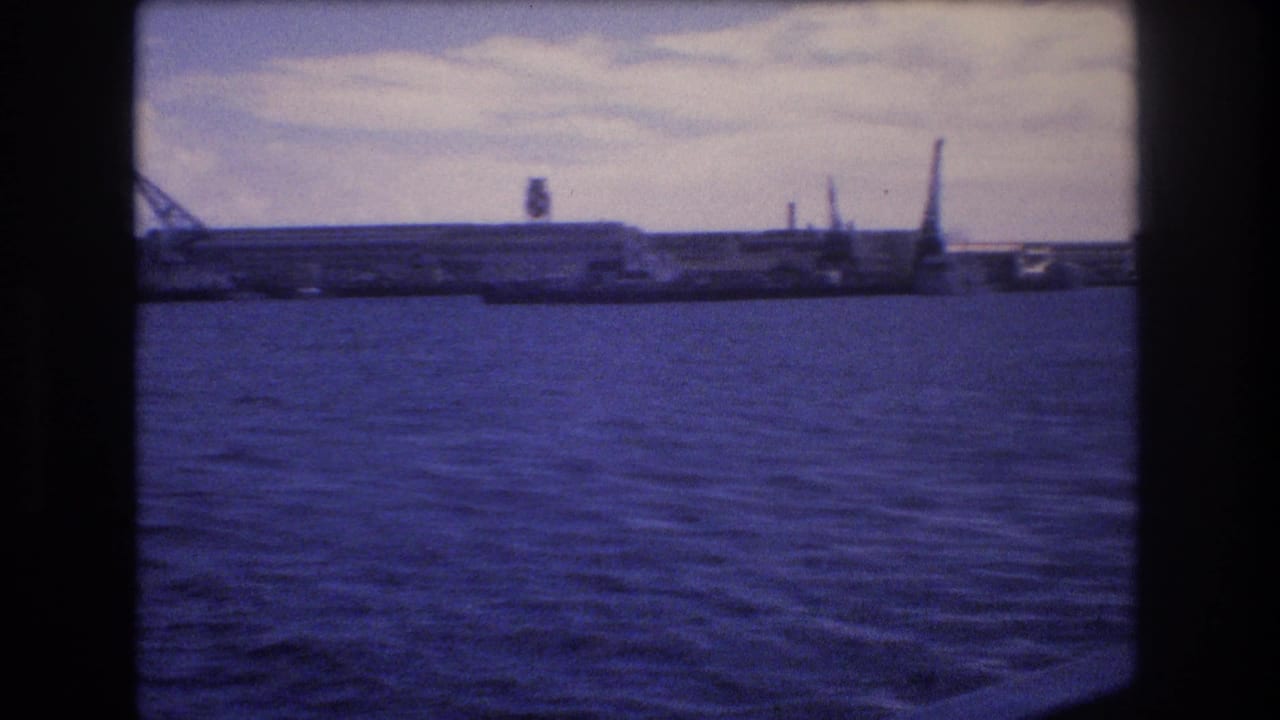 1977 Honolulu home-movie frame looking across blue water toward a distant industrial shoreline with cranes and low structures under a cloudy sky.