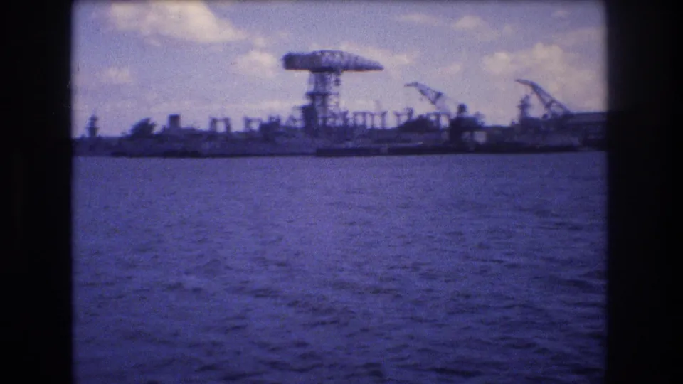 1977 Honolulu shoreline frame showing calm blue water, a prominent gantry-like structure, cranes, and low industrial forms in soft home-movie focus.
