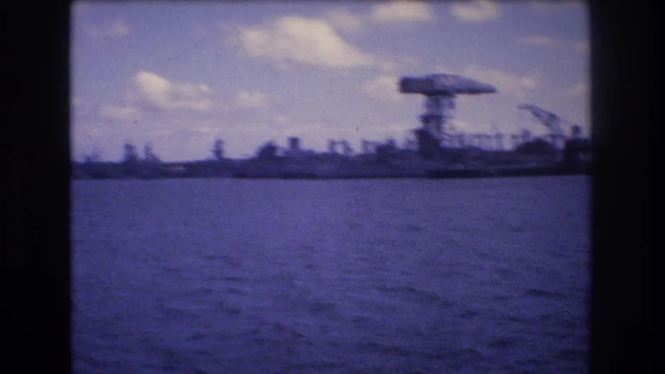 Late clip frame from the 1977 Honolulu harbor view, with a large gantry-like structure and cranes receding along the waterfront beyond blue water.