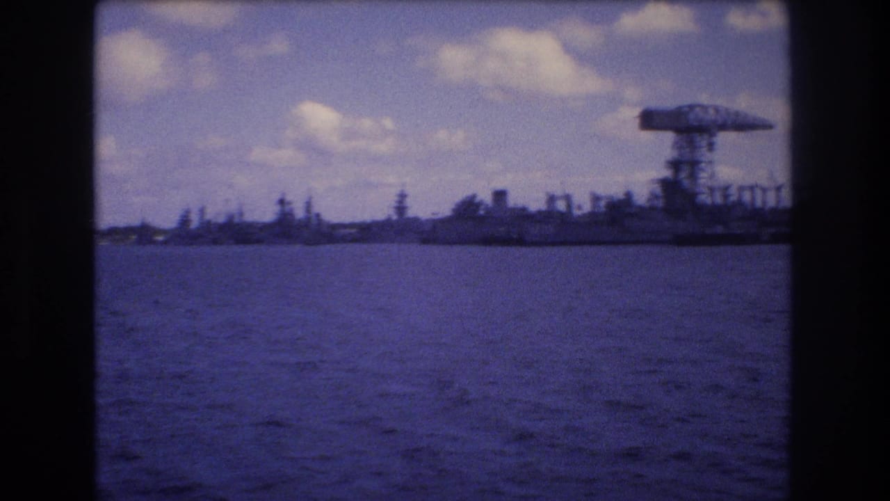 1977 Honolulu-area harbor view from a moving boat, showing blue water, a distant industrial shoreline, and a large crane or gantry against a cloudy sky.