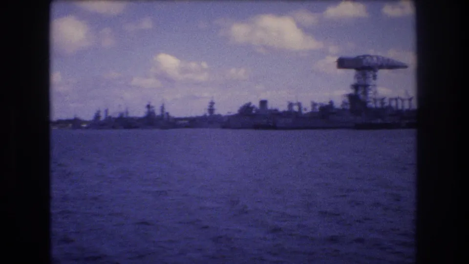 1977 Honolulu-area harbor view from a moving boat, showing blue water, a distant industrial shoreline, and a large crane or gantry against a cloudy sky.