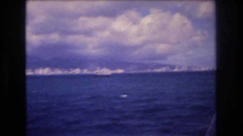 Boat-level view over dark blue water toward a distant Honolulu-area shoreline, with bright coastal forms beneath heavy clouds.