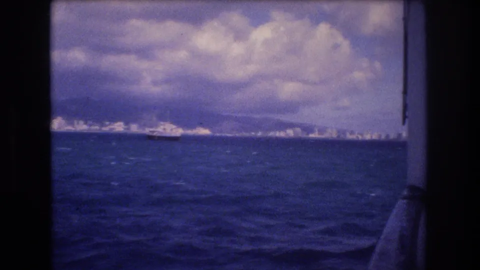 1977 Honolulu-area boat view with rough blue water in the foreground, a distant city shoreline, low clouds, and part of the filming boat visible at right.