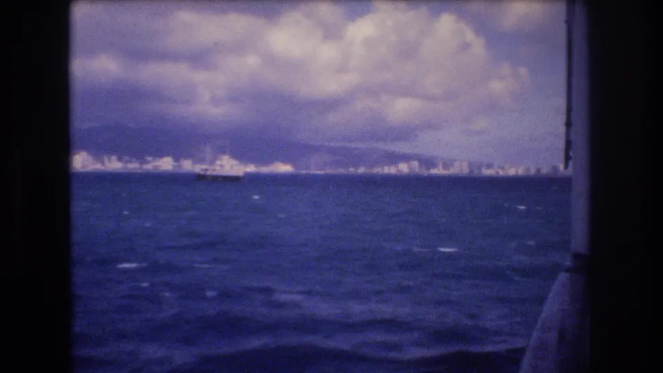 Wide 1977 Honolulu-area harbor view from a boat, showing choppy water, a small distant vessel, shoreline buildings, and a cloudy sky.