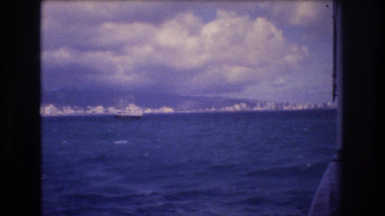 1977 8mm shipboard view approaching Honolulu, with deep blue water, cloudy sky, distant city skyline, mountains, and a large vessel offshore.