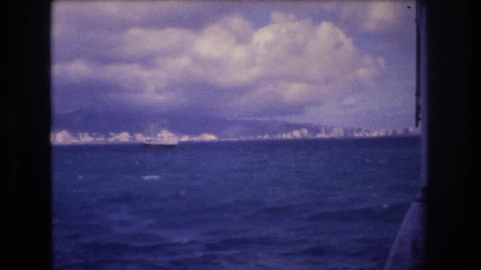 1977 8mm shipboard view approaching Honolulu, with deep blue water, cloudy sky, distant city skyline, mountains, and a large vessel offshore.
