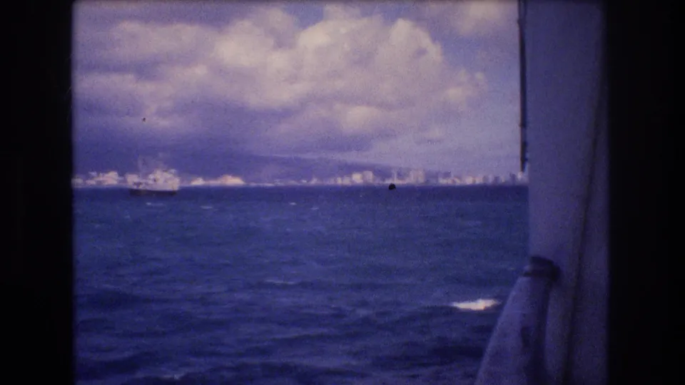 Shipboard 8mm view toward Honolulu in 1977, framed by dark onboard structure with ocean, clouds, distant skyline, mountains, and a vessel near the horizon.
