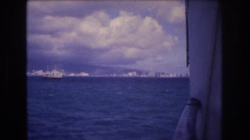 1977 home-movie frame from a ship nearing Honolulu, showing rough blue water, distant waterfront buildings, mountains under cloud, and ship hardware along the right edge.