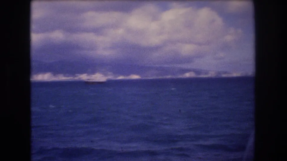 Final 8mm frame of the Honolulu approach, with dark water in the foreground, a distant coastal skyline, mountains under layered clouds, and part of the ship visible at right.