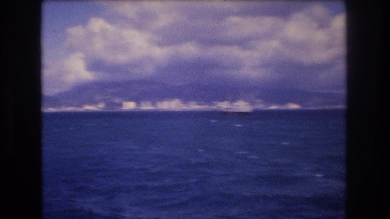 1977 8mm home-movie frame from offshore Honolulu showing blue ocean, distant coastal buildings, mountain ridges, and a dramatic cloud bank.