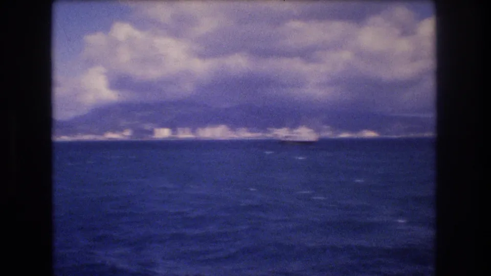 1977 8mm home-movie frame from offshore Honolulu showing blue ocean, distant coastal buildings, mountain ridges, and a dramatic cloud bank.