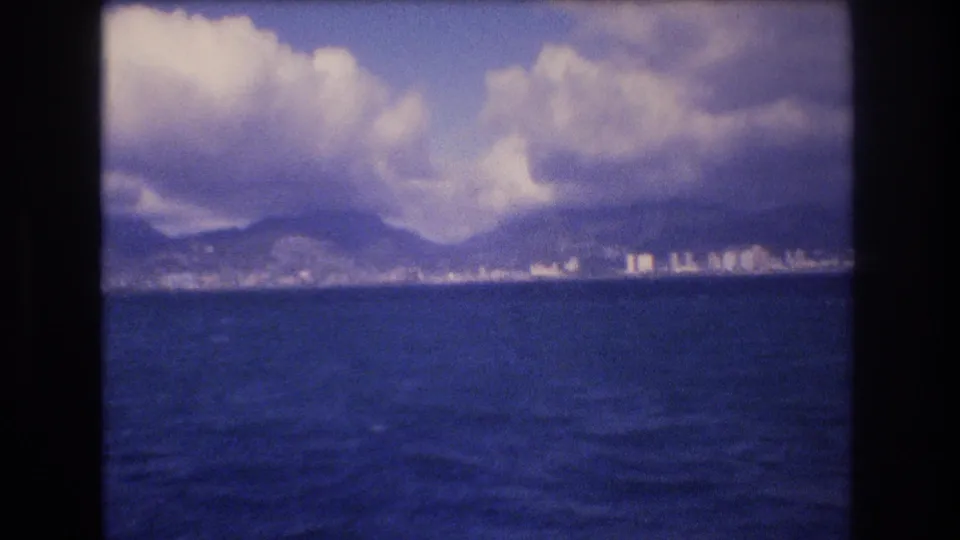 Offshore Honolulu view in 1977 8mm footage, with textured blue water in the foreground and distant buildings below cloud-covered mountains.