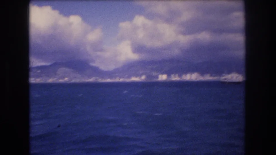 1977 Honolulu shoreline seen from a boat, with ocean filling the foreground and small bright buildings along the base of hazy mountains.