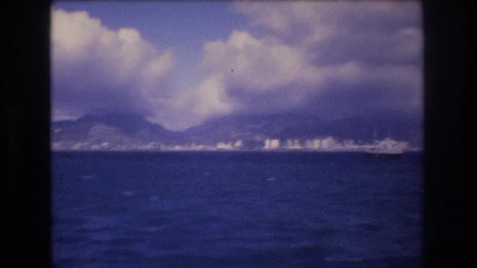 Final 1977 8mm frame of the Honolulu coast from offshore, showing rippled water, distant buildings, mountain slopes, and a small vessel or wake near the shoreline.